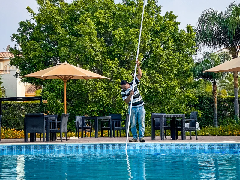 worker cleaning a pool with a cleaning tool with a yellow umbrella and black chairs and tables behind him